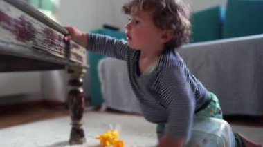 Meraklı çocuk oturma odasında masanın yanında durmak için yükseliyor, dengeyi ve erken vücut kontrolünü keşfederken yeni bir harekete odaklanıyor ve destek alanı algılıyor