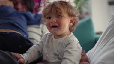 Yürüyen çocuk aile üyelerine yakın oturuyor ve neşeli bir ifadeyle kameraya doğru dönüyor. Bu an, rahatlamış bir ev ortamında sıcaklığı, eğlenceyi ve bağlantıyı iletir..