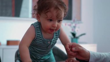 Bebek yüksek sandalyeden kalkıyor ve yoğun bir bakışla masanın üzerine eğiliyor, aşağıdaki yemek parçalarını gözlemliyor ve doğal bir merak ve aktif bir bağlılıkla alanı keşfediyor.