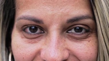 Kahverengi gözlü sarışın kadın. Göz kalemi çerçeveli. Kameraya bakarken nazikçe gülümsüyor.