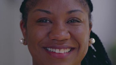 Gülümseyen Afro-Amerikalı kadın kameraya sıcak ve kendinden emin bir ifadeyle bakıyor. İnci küpeler takıyor ve saçlarını örüyor.