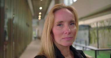 İş kıyafetleri içinde kendinden emin bir kadın doğrudan kameraya bakıyor. Modern ofis koridorunda profesyonel portre için oluşturulmuş bir ifade.