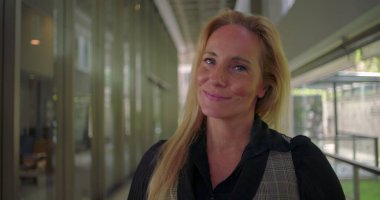 Modern koridorda kurnazca gülümseyen bir kadına yakın dur. Profesyonel çevrede kamerayla göz teması kurarken sıcak ve sessiz bir güven yayar.