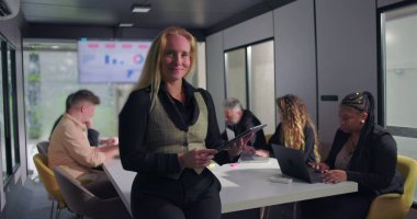 Ofis toplantısı sırasında konferans masasının başında duran neşeli kadın yönetici, verimli ve olumlu bir ortamda arka planda çalışan takım.