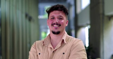 Modern ofis koridorunda gülümseyen, kısa saçlı ve sakallı, kollarını kavuşturmuş, özgüven ve samimiyet saçan adamın yakın çekimi.