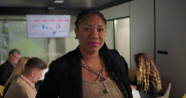 Kendine güvenen Afrikalı Amerikalı iş kadını ofisteki kameraya bakıyor, konferans masasının yanında dururken takım dizüstü bilgisayarlar ve veri tablosu üzerinde çalışıyor.