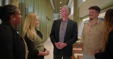 Şirket yöneticisi modern ofis koridorunda çeşitli takımlara sesleniyor, bir grup profesyonel gayri resmi toplantı sırasında iş yerinde sohbet ediyorlar.