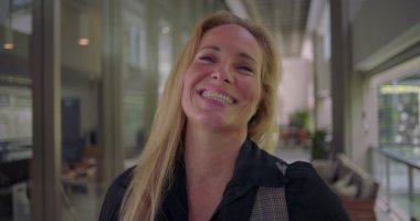 Kameraya hafifçe gülümseyen bir kadın, koridorda dururken kibarlık, profesyonellik ve kendine güven yansıtıyor.