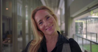 Kameraya hafifçe gülümseyen bir kadın, koridorda dururken kibarlık, profesyonellik ve kendine güven yansıtıyor.