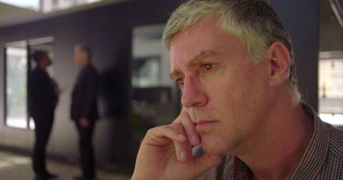 Orta yaşlı, düşünceli bir yüz ifadesine sahip, elleri çenesinde dinlenen, sessizlikte ileriye bakan, arka planda bulanık meslektaşlarıyla ofis ortamında oturan bir adama yakın durun.