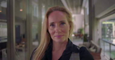 Sakin ve açık bir ifadeyle kameraya bakan, koridorda hareketsiz duran, kendine güveni ve sessizliği temsil eden kadına yakın dur.