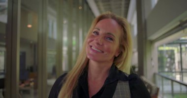 Modern koridorda kurnazca gülümseyen bir kadına yakın dur. Profesyonel çevrede kamerayla göz teması kurarken sıcak ve sessiz bir güven yayar.