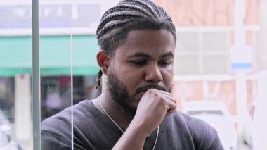 Saçları örgülü ve sakallı genç siyahi adam sessiz ve düşünceli bir ifadeyle aşağıya bakıyor.