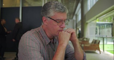 Orta yaşlı bir adam modern ofis ortamında masada oturuyor, elleri ağıza yakın, endişeli ve dalgın bir halde aşağıya bakıyor, kaygı ve duygusal karmaşa gösteriyor.