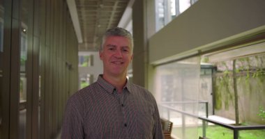 Orta yaşlı bir adam koridorda duruyor. Yumuşak bir gülümsemeyle ofisin iş yeri koridorunda geniş pencereli rahatlamış bir profesyonel ifadeyle.