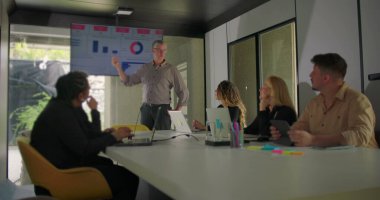 Çeşitli profesyoneller modern konferans odasında doğal ışık ile sunum izlerken, yönetici ekranda veri sunar 