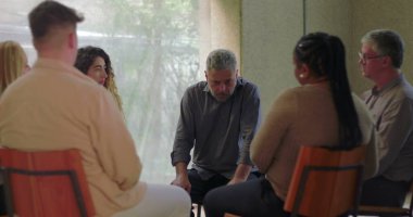 Adam grup terapisi sırasında konuşur, öne doğru eğilir ve sessiz odada diğerleriyle doğal ışık ve duygusal odaklanma dolu destek çemberine girer.