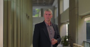 Orta yaşlı bir adam modern ofis koridorunda duruyor ceketini ayarlıyor, kendine güvenen ve parlak profesyonel çevrede yaşıyor.