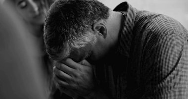 Bir erkek terapi seansında duygusal tepki verir, gözleri kapalı ve ağzı gergin başka bir katılımcı omzunda destekleyici bir elle şefkatli bir an yaşar. 