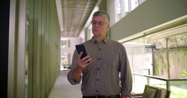 Orta yaşlı bir adam modern ofis koridorunda cep telefonu kullanarak ekranı tarıyor ve iş ortamında mobil cihazla etkileşime geçiyor.