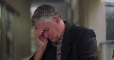 Orta yaşlı bir adam ofis odasında oturuyor, elini yüzünü kapatıyor, ev ortamında duygusal acı ve pişmanlık ifade ediyor. 