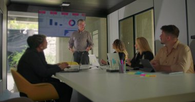 Çeşitli profesyoneller modern konferans odasında doğal ışık ile sunum izlerken, yönetici ekranda veri sunar 