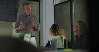 Kadın meslektaşlar dijital ekrandaki veri tablolarına bakarken, erkek profesyoneller konferans odasında grup sunumu yapıyor. 