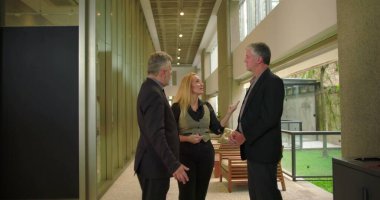 Modern ofis koridorunda konuşan bir grup iş arkadaşı, gayriresmi toplantı sırasında gevşek iş yeri sohbetine girişen profesyonel takım. 