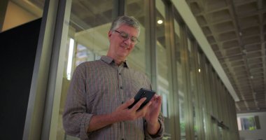 Ofis koridorunda cep telefonu kullanırken gülümseyen orta yaşlı bir adam aydınlık, modern iş çevrelerinde neşeli ve rahat bir ifadeyle ekrana bakıyor.