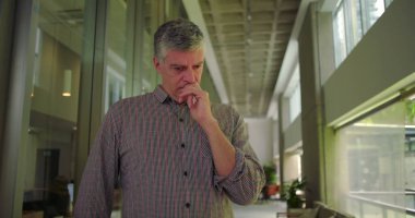 Orta yaşlı bir adam ofis koridorunda elleri ağızlarına yakın duruyor endişeli ifadeye bakıyor profesyonel settin 'deki endişe ve endişe dolu stresi iletiyor.