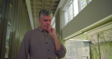 Orta yaşlı bir adam ofis koridorunda, iki elini de yan yana tutarak aşağıya bakıyor. Şirket çevresindeki duygusal endişe ve endişeyi gösteriyor.