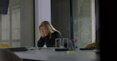Kadın, modern ofiste masa başında otururken yüzünü iki eliyle kapatıyor. Sessiz bir şirket ortamında stres ya da duygusal hayal kırıklığını ifade ediyor.