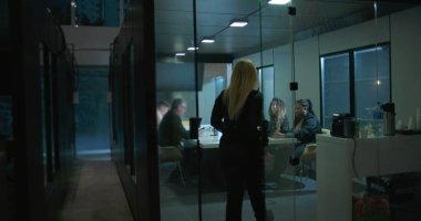 Meslektaşlar modern kurumsal çalışma ortamında geç saatlerde ofis toplantılarında dizüstü bilgisayarlarla masaya otururken cam toplantı odasına giren kadın yönetici.
