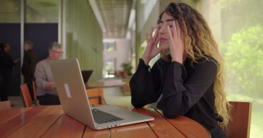 Hüsrana uğramış bir kadın, modern ofis salonunda dizüstü bilgisayarda otururken geriye yaslanıyor ve elleriyle yüzünü bastırıyor. Bu da iş stresini gösteriyor.