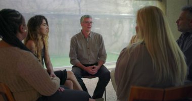 Orta yaşlı bir adam terapi grubu oturumunda duygusal ifadelerini paylaşıyor. Katılımcılar hassas destek ve iyileşme anlarında dikkatle dinliyorlar.