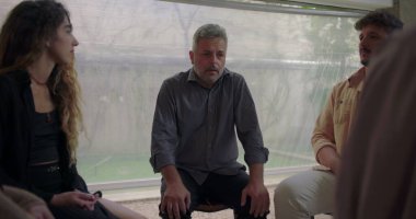 Duygusal grup terapisinde kişisel mücadeleyi ortaya koyan, katılımcılar güvenli iyileşme alanında empatiyle dinlerken, aciliyet ve kırılganlıkla konuşan bir adam.