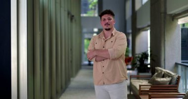 Ofis koridorunda kolları çapraz duran, kendine güvenen, modern çalışma alanında kendine güvenen, doğal ışık alan bir adam.