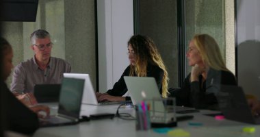 Modern çalışma ortamında masanın etrafında oturan çeşitli meslektaşlarla sessiz ofis toplantısı sırasında dizüstü bilgisayarlar üzerinde çalışan odaklanmış iş ekibi.