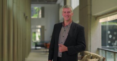 Modern ofis koridorunda gülümseyen orta yaşlı bir adam, kendine güvenen ve parlak profesyonel bir çalışma alanında yaklaşımlı görünüyor.