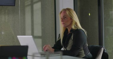 Modern cam duvarlar ve minimum dikkat dağınıklığıyla sessiz ofis konferans odasında tek başına çalışırken dizüstü bilgisayara konsantre olan profesyonel bir kadın.