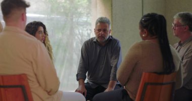 Adam grup terapisi sırasında konuşur, öne doğru eğilir ve sessiz odada diğerleriyle doğal ışık ve duygusal odaklanma dolu destek çemberine girer.