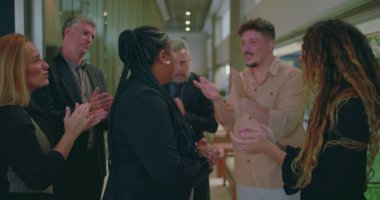 Kadın takım üyesi ofis koridorunda sıcak bir alkış alıyor, meslektaşları birlikte destek veriyorlar ve profesyonel dönüm noktasını kutluyorlar.