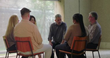 Adam grup terapisi sırasında konuşur, öne doğru eğilir ve sessiz odada diğerleriyle doğal ışık ve duygusal odaklanma dolu destek çemberine girer.