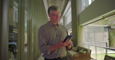 Orta yaşlı bir adam ofis koridorunda gülümserken cep telefonu gevşetici ifadeler kullanıyor. Parlak modern çalışma plağındaki mobil cihazlara bağlanıyor.