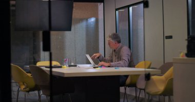 Erkek profesyonel, akşam ışığında ofis masasında tek başına çalışır, odaklanmış ifadeyle, belgelerin ve renkli ofislerin ortasında dizüstü bilgisayarda yazar.