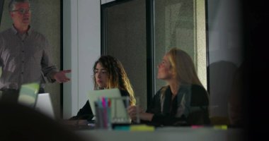 Kadın meslektaşlar dijital ekrandaki veri tablolarına bakarken, erkek profesyoneller konferans odasında grup sunumu yapıyor.