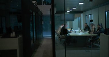 Şirketin gece vardiyası sırasında cam toplantı odasında laptoplar ve belgeler masaya serilerek geç saatlere kadar çalışan çeşitli takımların geniş görüşü.