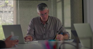 Olgun iş adamı, elinde kalem tutarken ve modern konferans masasında bilgileri incelerken sessiz ofis toplantısı sırasında dizüstü bilgisayarda dikkatle çalışıyor.