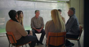 Orta yaşlı bir adam terapi grubu oturumunda duygusal ifadelerini paylaşıyor. Katılımcılar hassas destek ve iyileşme anlarında dikkatle dinliyorlar.