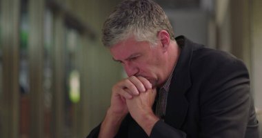 Ofis salonunda elleri sımsıkı bağlı orta yaşlı bir adam, derin düşüncelere dalmış ve duygusal mücadele belirtileri gösteriyor.